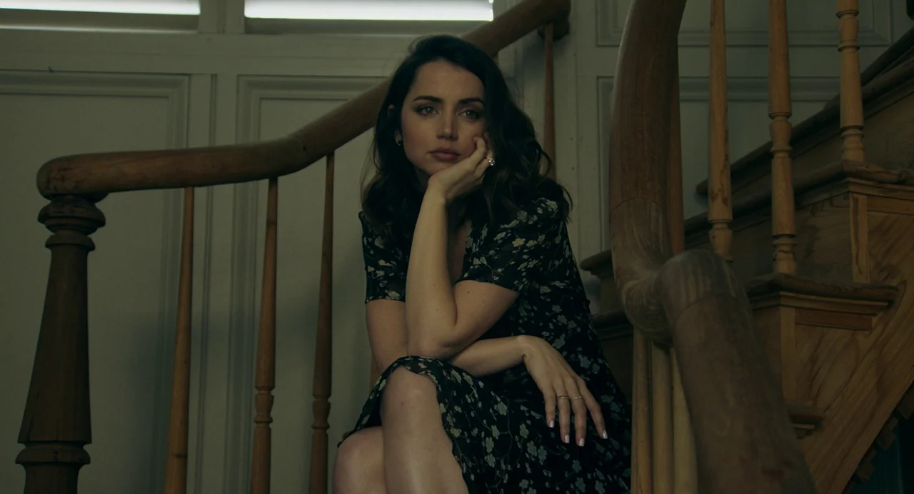 Deep Water (2022) movie still: medium — A woman sits on the landing of a wooden staircase inside a house. She rests her chin on her hand while…