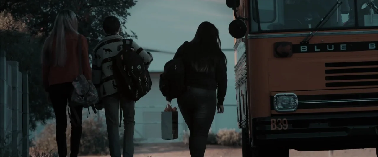 Anomaly (2006) movie still: fullbody — A rural bus stop features a large orange school bus parked on the side. Three students walk toward the open…