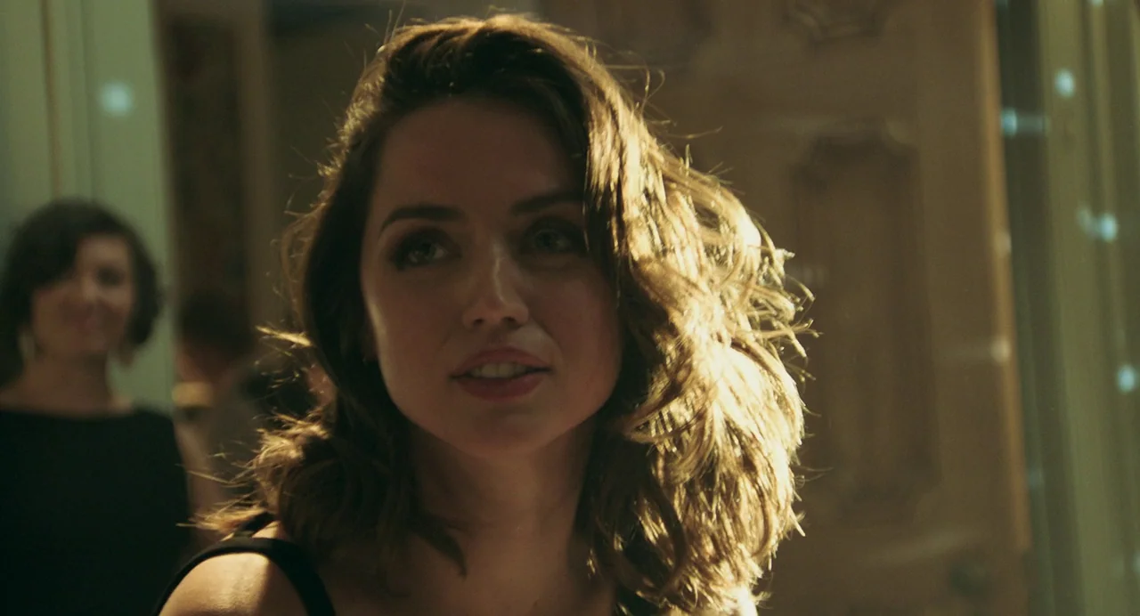 Deep Water (2022) movie still: closeup — A dimly lit indoor gathering with blurred shapes and figures in the background. A woman with…