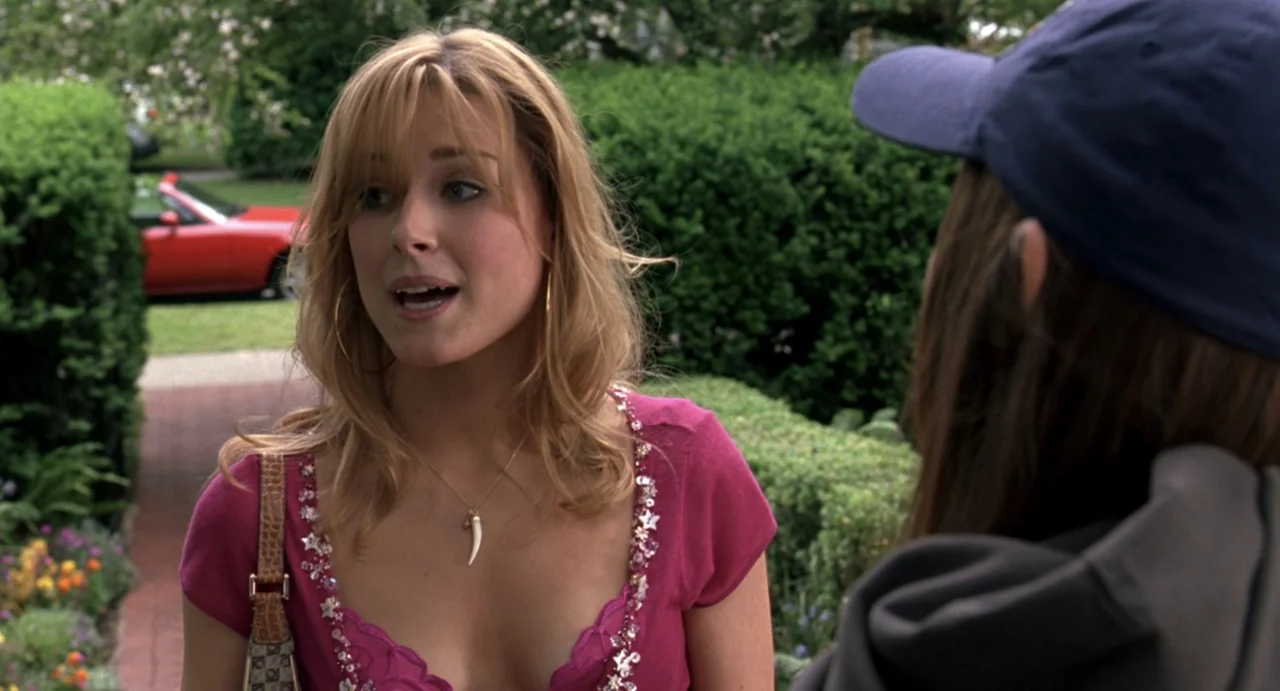 She's the Man (2006) movie still: medium — A residential driveway is bordered by manicured green hedges and colorful flowers. A blonde woman speaks…