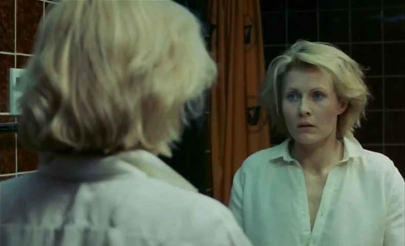 No End (1985) movie still: medium — A bathroom with dark tiled walls and an orange curtain hanging in the background. A woman in a white shirt…