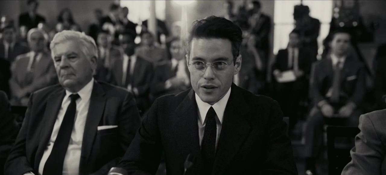Oppenheimer (2023) movie still: medium — A crowded formal room with attendees sitting in rows behind the main subjects. A younger man in wire-rimmed…