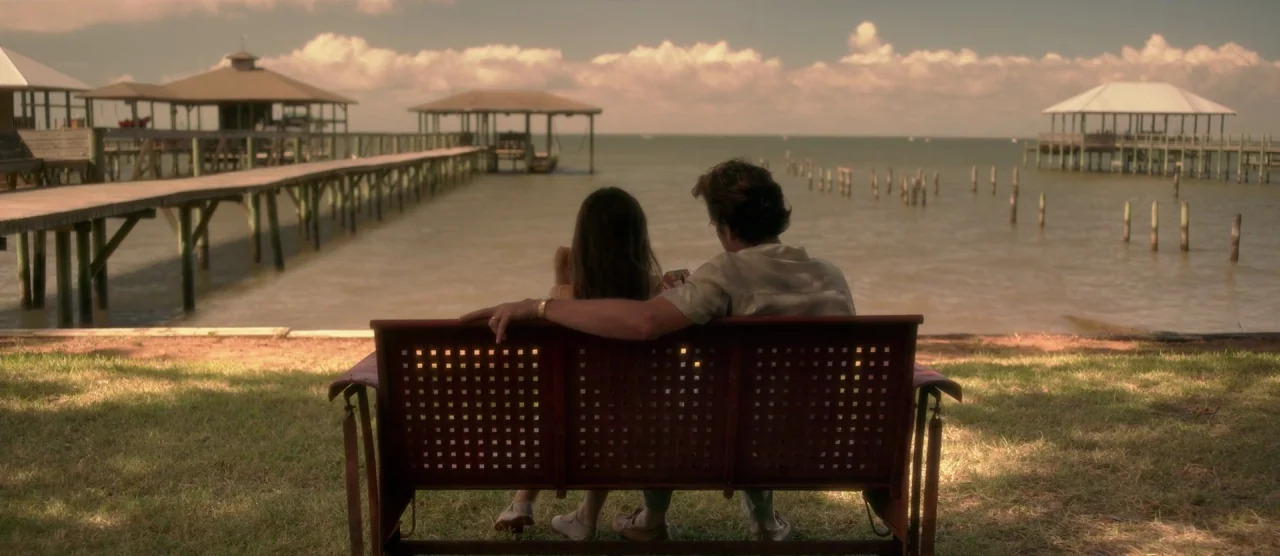 Gerald’s Game (2017) movie still: fullbody — A grassy bank overlooks a large body of water with multiple wooden docks. A young man and woman sit…