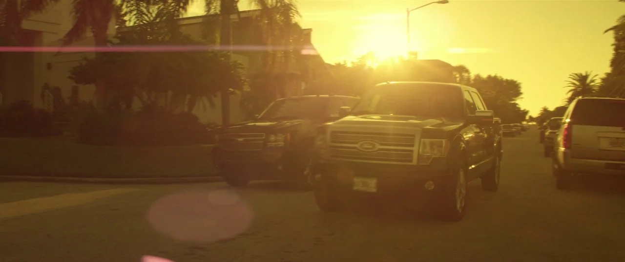 Magic Mike (2012) movie still: wide — A quiet suburban street is lined with parked pickup trucks during a sunset. The low sun creates a strong…
