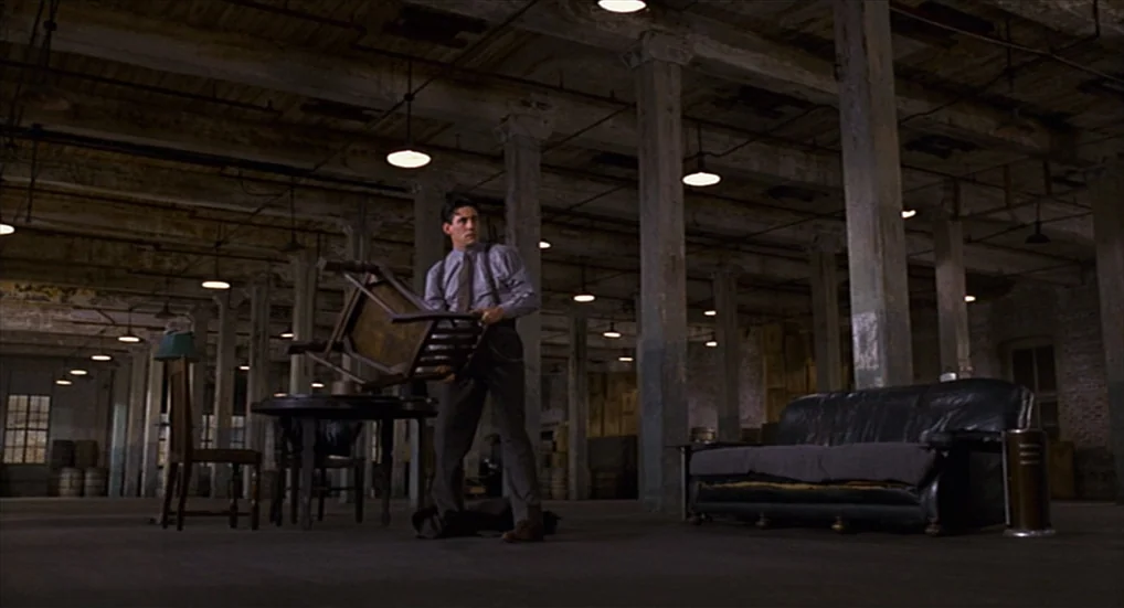Miller’s Crossing (1990) movie still: wide — A vast, empty industrial interior features concrete pillars and rows of hanging light fixtures. A man…