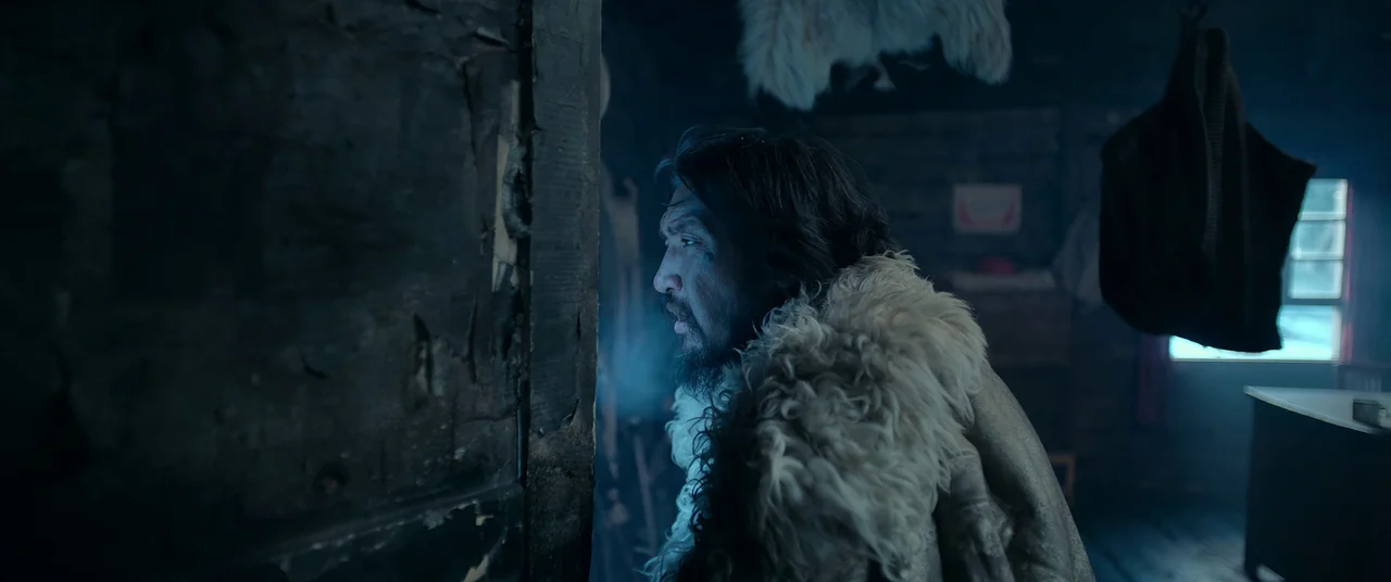 One And Four (2021) movie still: medium — A dimly lit interior room features dark wooden walls and fur-lined textures. A bearded man with a fur coat…