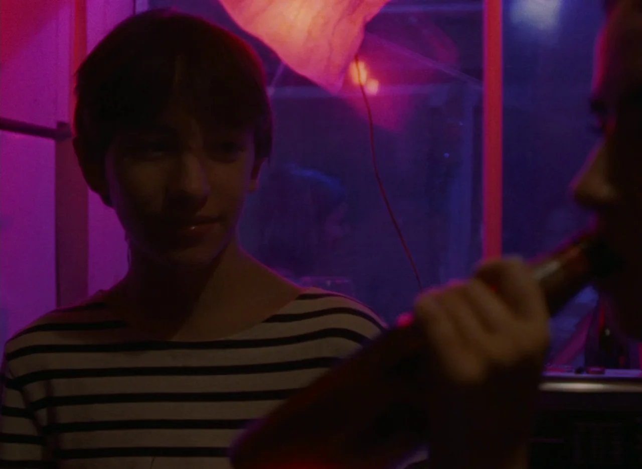 Falcon Lake (2022) movie still: medium — A dimly lit room with neon purple and magenta light filling the space. A teenager wearing a striped shirt…