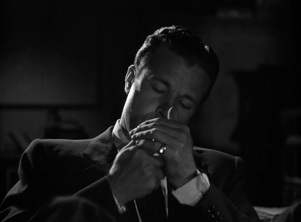 Murder, My Sweet (1944) movie still: closeup — A man stands in a dimly lit, nondescript room shrouded in shadow. He cups his hands together while…