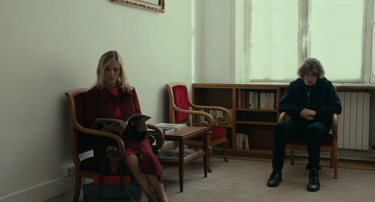 Last Summer (2023) movie still: wide — A quiet room features white walls, wooden chairs, and a small library shelf. A woman reads a magazine while a…