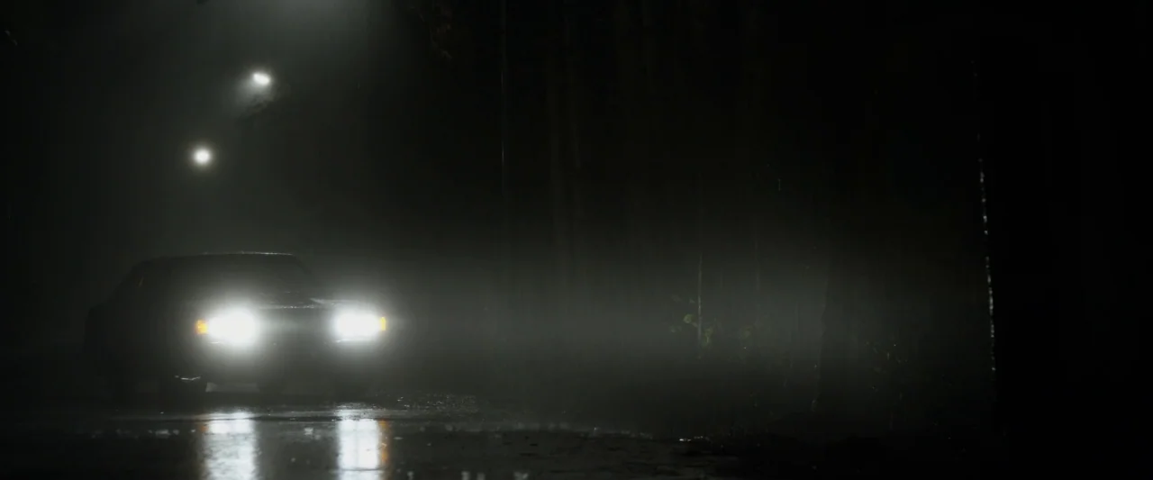 The Monster (2016) movie still: wide — A dark, wet road at night is surrounded by tall, obscured trees in heavy mist. A car with its headlights…