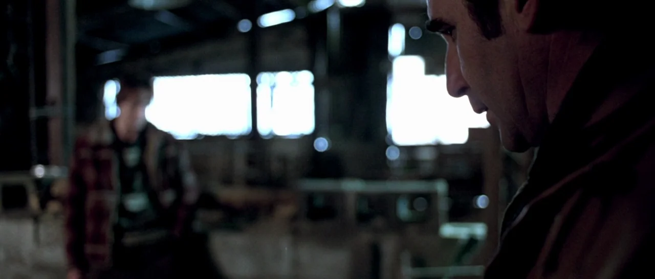 Isolation (2005) movie still: medium — A dark industrial building has large window openings letting in intense bright daylight. Two men stand in the…