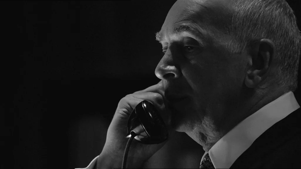 Good Night And Good Luck (2005) movie still: closeup — An elderly man is framed in a dark office environment. He holds a black vintage telephone…