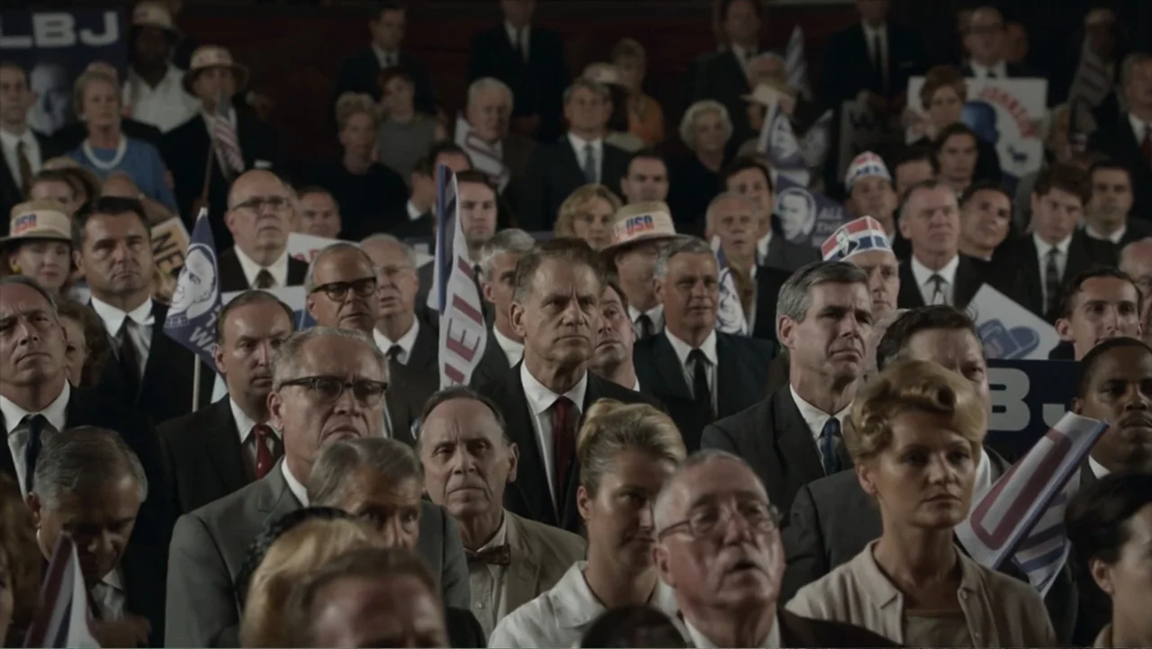 All the Way (2016) movie still: wide — A dense indoor political rally filled with rows of spectators in formal 1960s attire. The crowd faces…