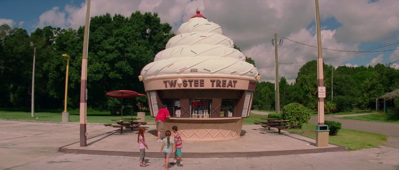 The Florida Project (2017) movie still: wide — An ice cream stand shaped like a giant cupcake sits in a parking lot. A man and three children stand…