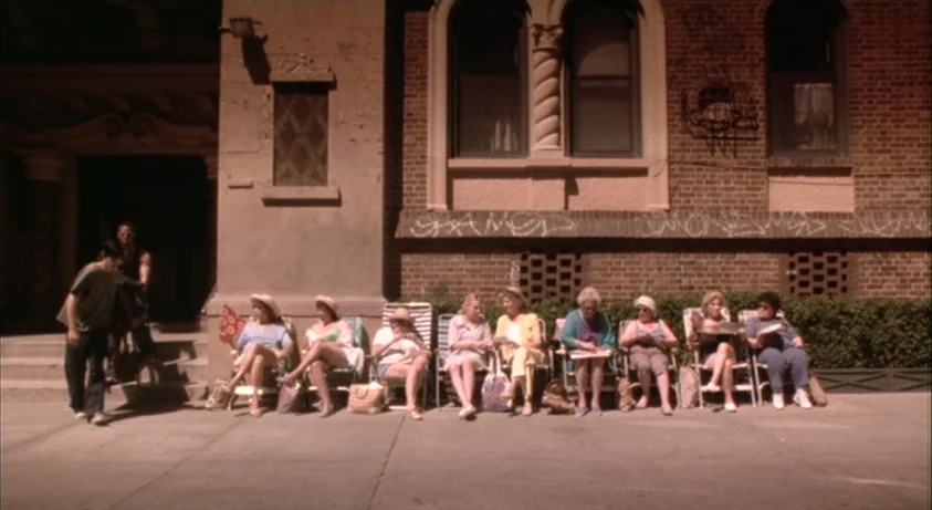 Requiem For A Dream (2000) movie still: wide — A line of eight women sit in individual folding lawn chairs against a brick building. The women watch…