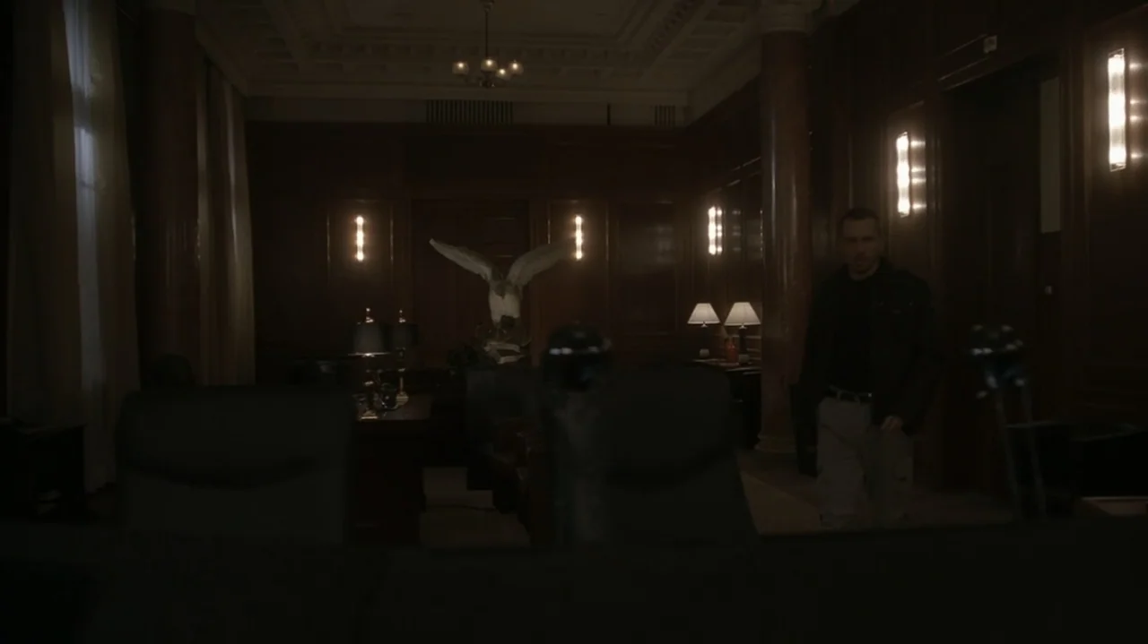 The Shepherd (2023) movie still: medium — A wood-paneled executive office with dark furniture and a large bird sculpture center-back. A man in a…