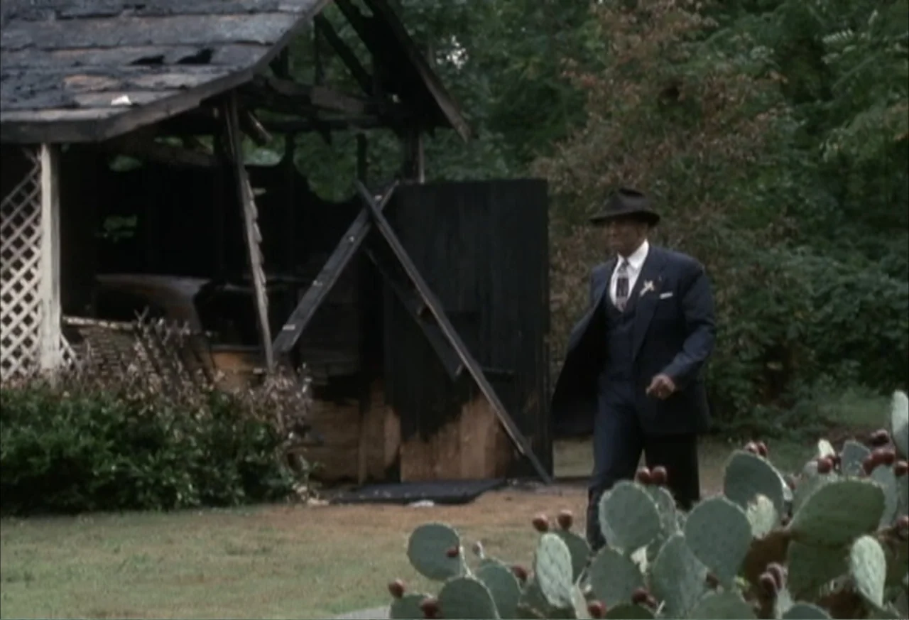 Boycott (2001) movie still: fullbody — A man walks past the charred remains of a wooden outbuilding in a field. Large prickly pear cacti occupy the…