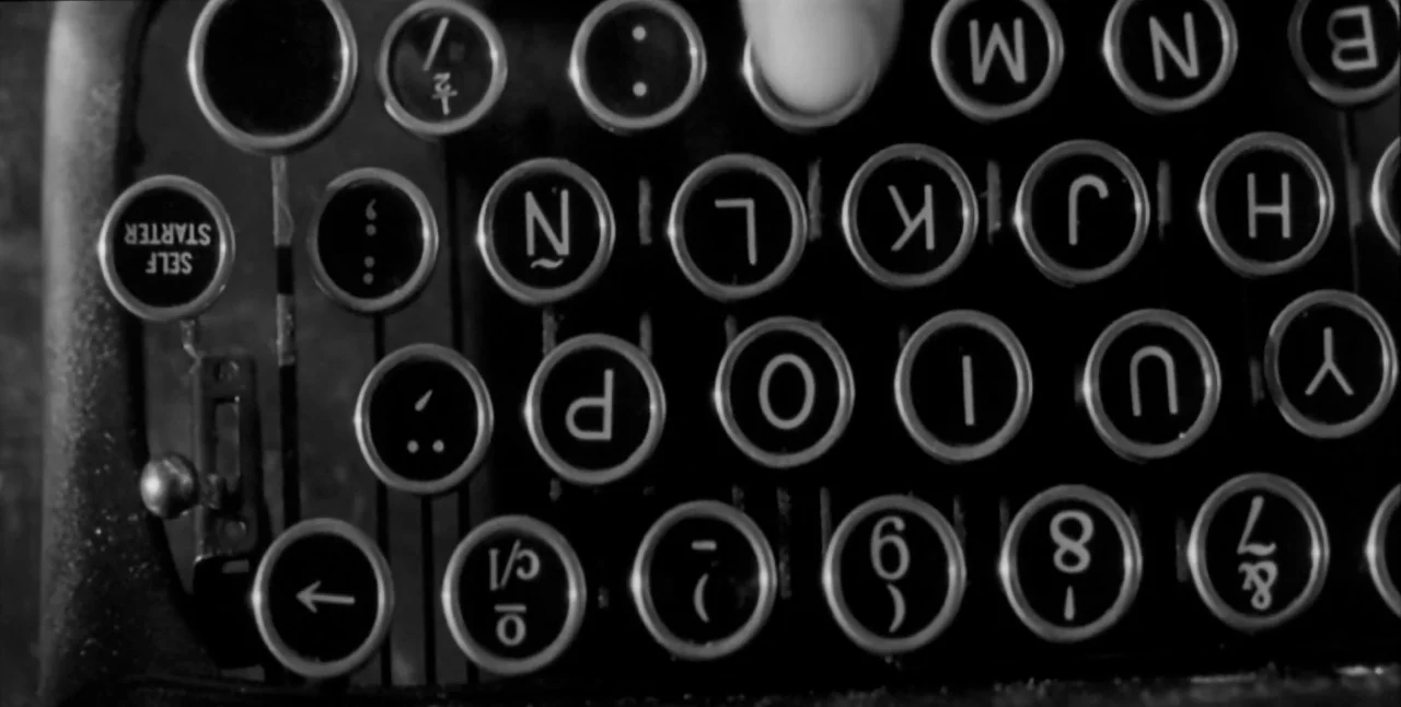The Aerial (2007) movie still: closeup — A close-up view of vintage typewriter keys arranged in a specific keyboard layout. One finger presses on a…