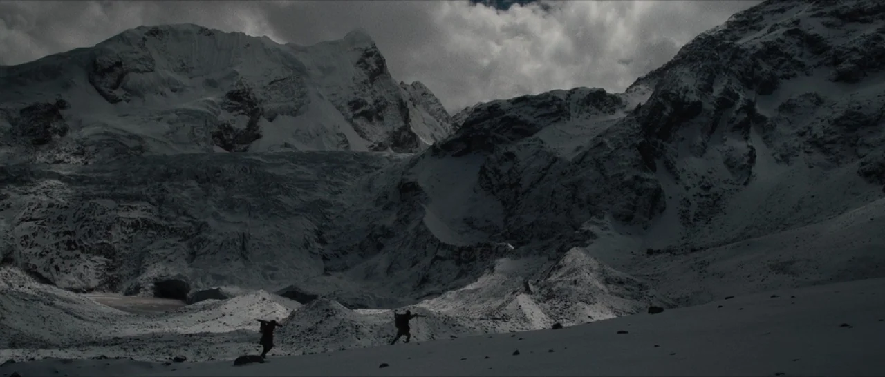 The Lost City of Z (2016) movie still: wide — A vast, rugged mountain range features snow-covered peaks and dark, rocky, glaciated slopes. Two tiny…