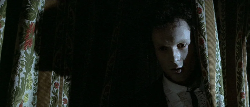 Whity (1971) movie still: closeup — A dark space framed by floral patterned fabric curtains on either side. A man with pale skin watches from behind…