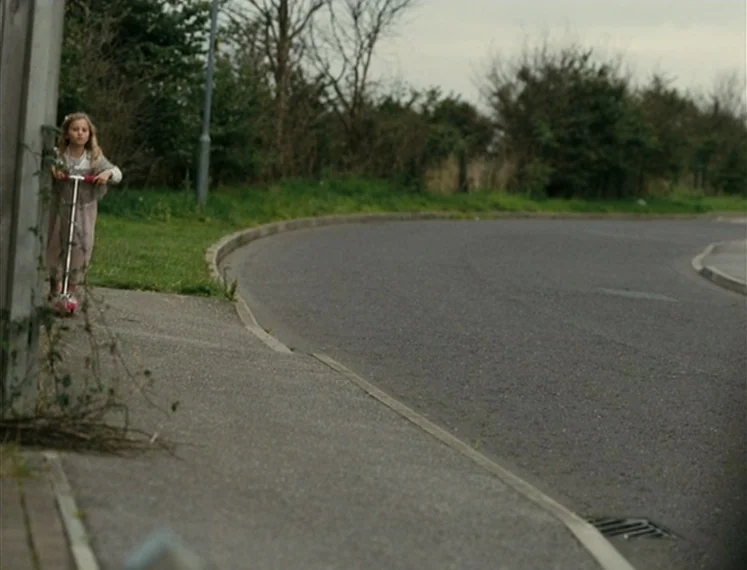 Fish Tank (2009) movie still: wide — A suburban road curves through a sparse neighborhood under an overcast gray sky. A young girl stands on a small…
