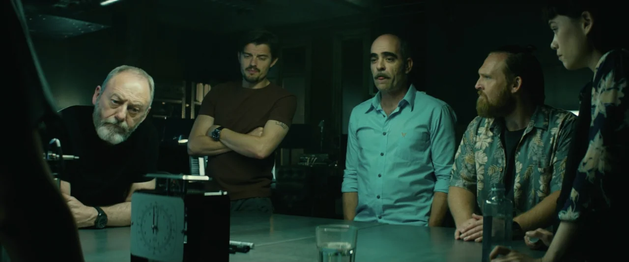 The Vault (2021) movie still: medium — A dimly lit room contains a metallic table where five individuals congregate. The group observes a small…