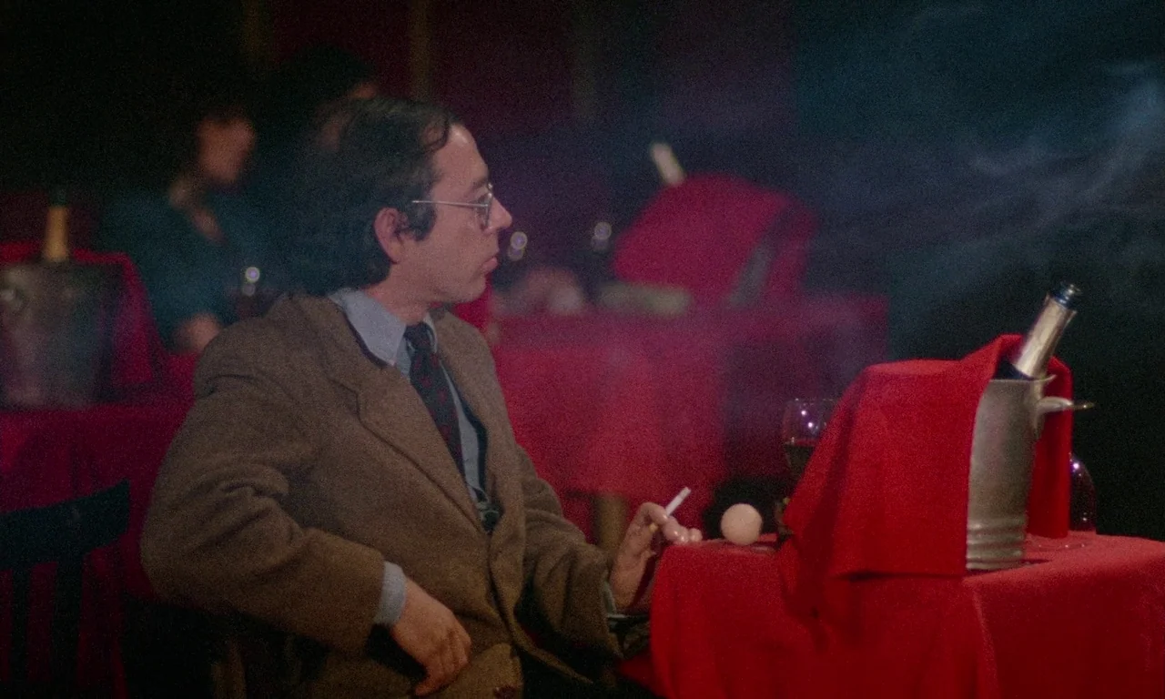 The Margin (1976) movie still: medium — A dimly lit room features tables covered with vibrant red cloths in the dark. A man in a brown blazer sits…