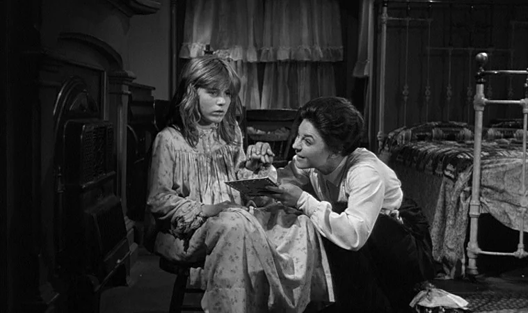 The Miracle Worker (1962) movie still: medium — A dark, furnished room contains a child in a nightgown and a seated woman. The woman presents a…