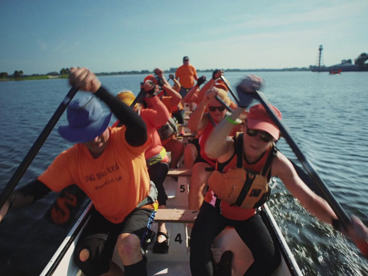 Some Kind Of Heaven (2020) movie still: medium — A group of people paddle a long narrow boat across a wide lake. Several rows of rowers in matching…