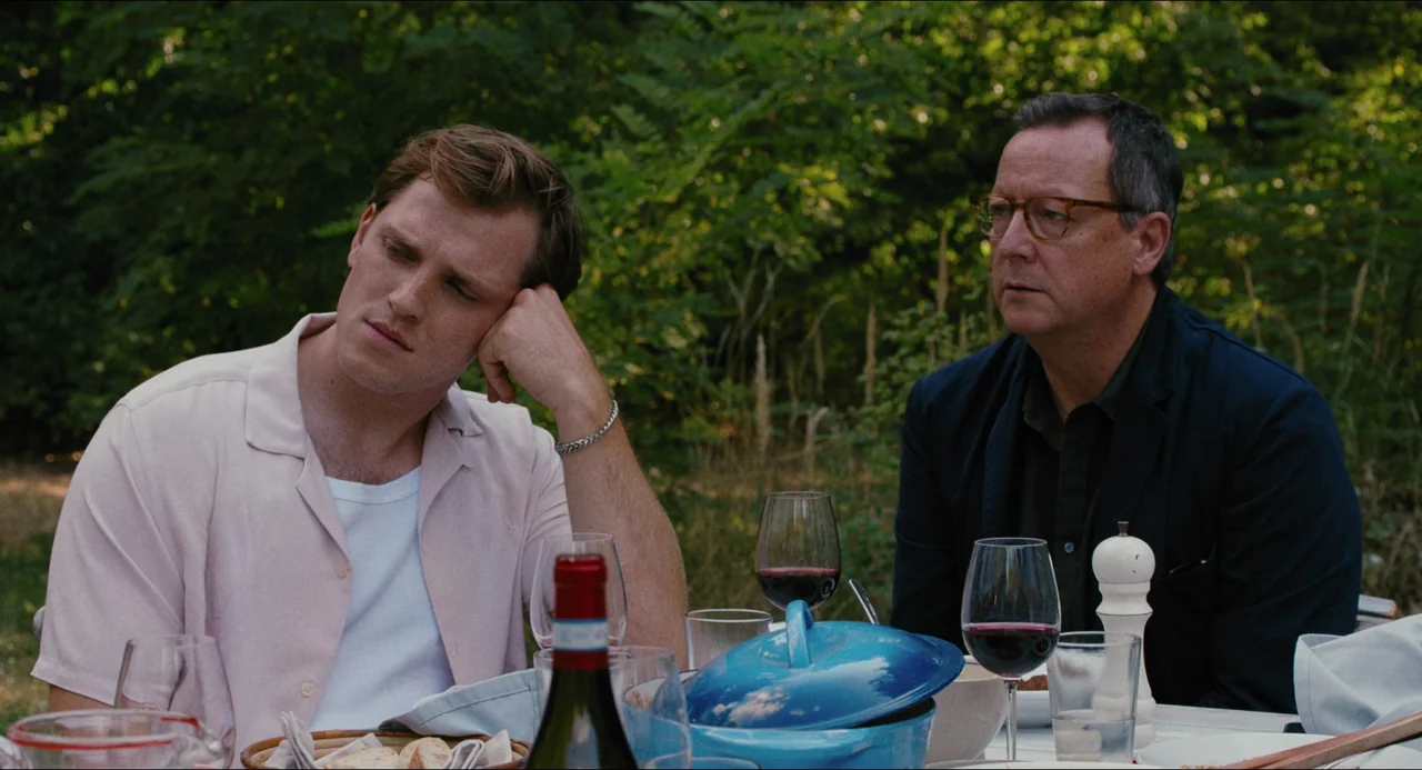 Afire (2023) movie still: medium — An outdoor dining setting features a table set with wine glasses and cookware. Two men sit together amidst a…