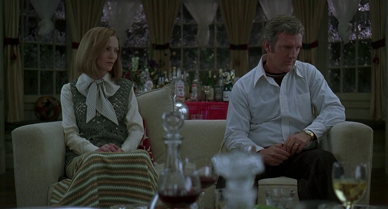 The Ice Storm (1997) movie still: medium — A 1970s sunroom features a beige sofa and a rear bar with liquor bottles. The man and woman sit upright…