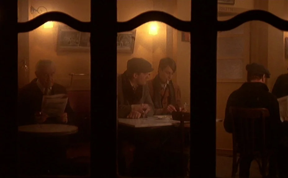 The Last Metro (1980) movie still: wide — An interior view of a dimly lit, vintage cafe seen through a window frame. Patrons sit at individual…