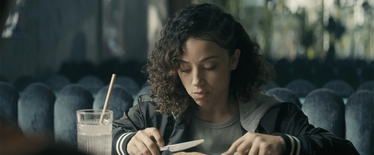 Perfect Addiction (2023) movie still: medium — A diner booth featuring grey cushioned seating and a glass of water with a straw. A young woman…