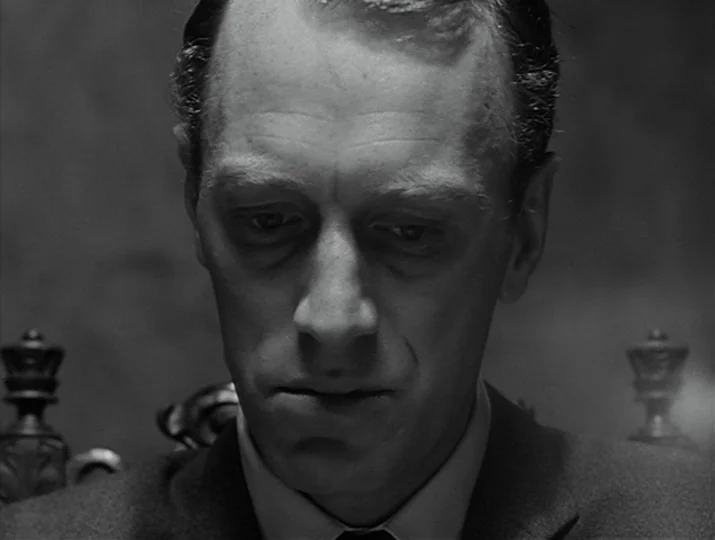 Hour of the Wolf (1968) movie still: closeup — A man in a suit sits in a room with a blurred wooden chair. He looks downward with a fixed expression.