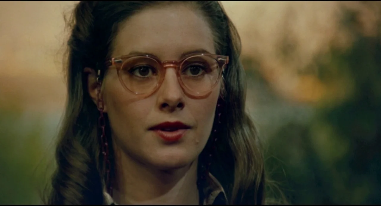 Olivia (1983) movie still: closeup — A woman stands outdoors in a nature setting during golden hour. She wears glasses with a red chain and looks…