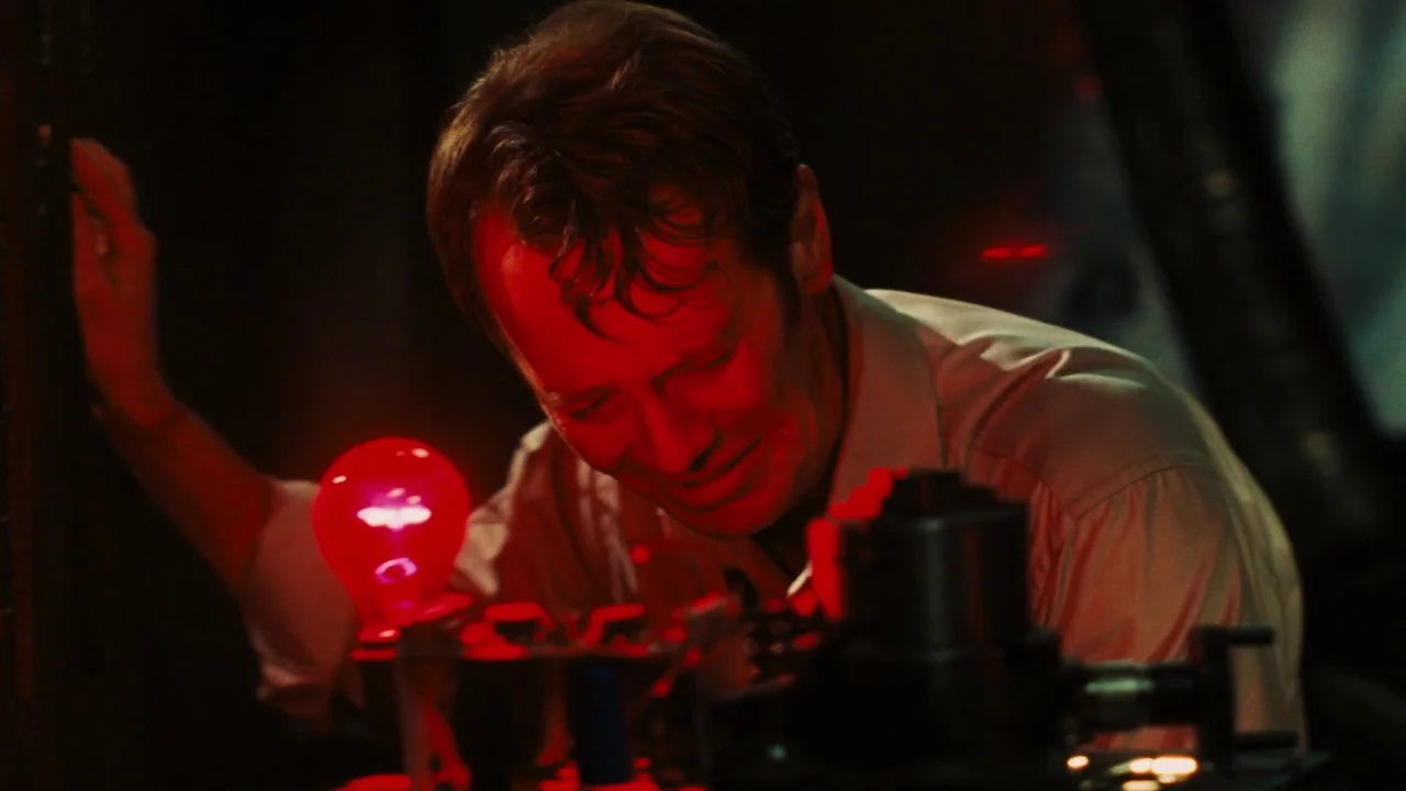Saw VI movie still: medium — A dark, cramped space filled with mechanical equipment and a single glowing red lamp. A man leans over a workbench,…