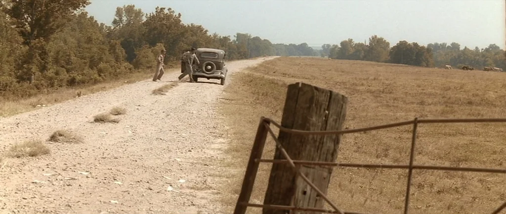 O Brother, Where Art Thou? (2000) movie still: wide — An unpaved road runs through a dry field flanked by trees under a haze. Two men stand beside…