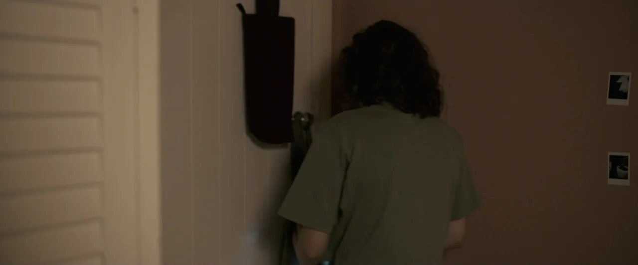 Talk to Me (2023) movie still: medium — A quiet bedroom corner features a closed door and beige walls with photos. A young woman with dark wavy hair…