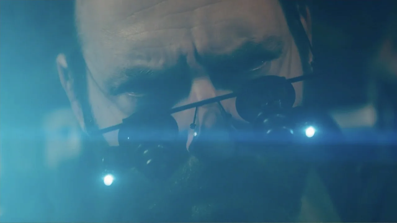 Robot Overlords (2015) movie still: closeup — A dimly lit room focused on a man wearing professional medical head-mounted surgical magnifiers. The…