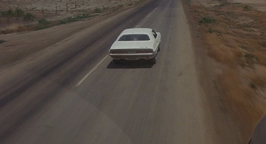 Vanishing Point (1971) movie still: wide — An arid, unpaved desert landscape surrounds a long, straight stretch of asphalt road. A white vintage…