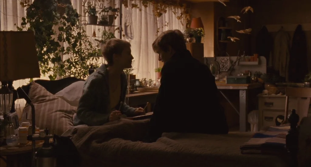 Restless (2011) movie still: medium — A cluttered room features a bed positioned in front of a window with sheer curtains. Two young people sit…