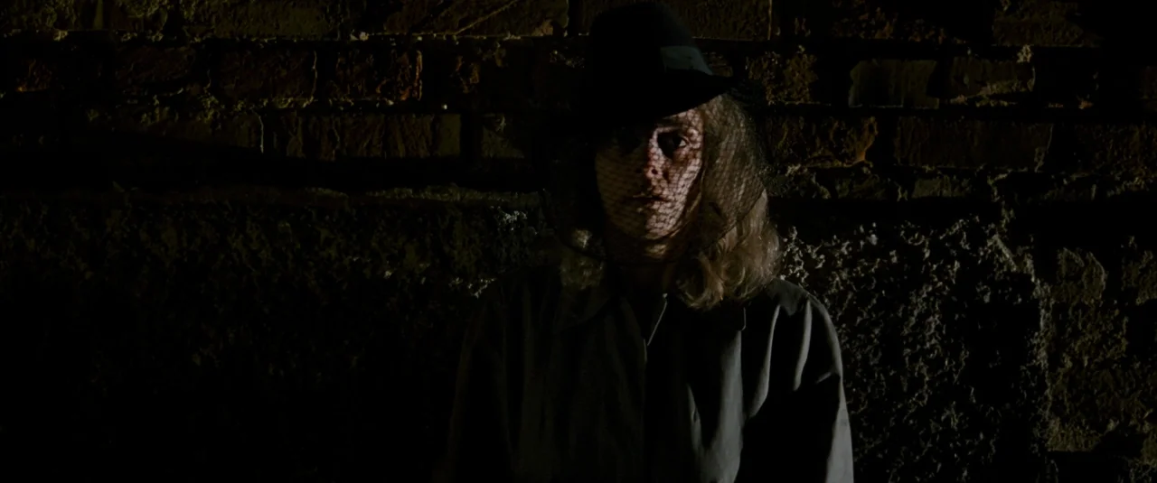 Phoenix (2014) movie still: medium — A woman stands in front of a dark, weathered brick wall at night. She is dressed in a trench coat and a veiled…