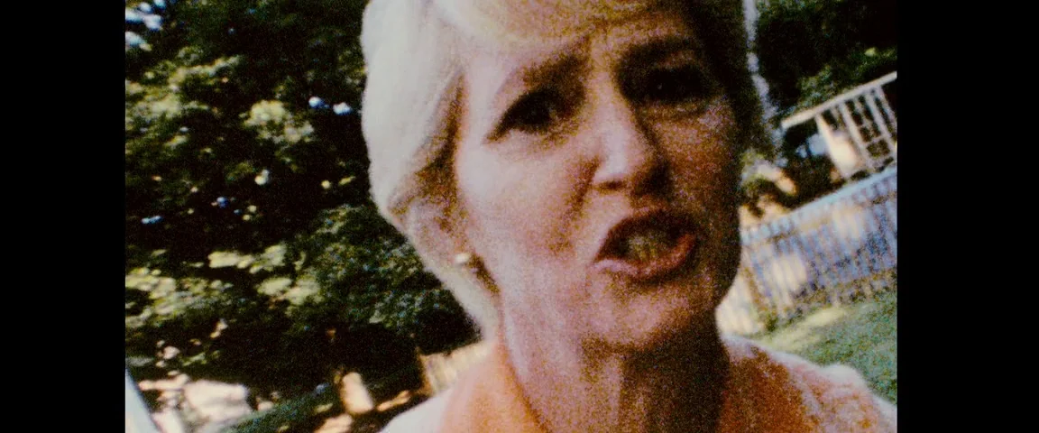 The Fighter (2010) movie still: closeup — An older woman stands in an outdoor setting with leafy green trees and fencing. She faces the camera…