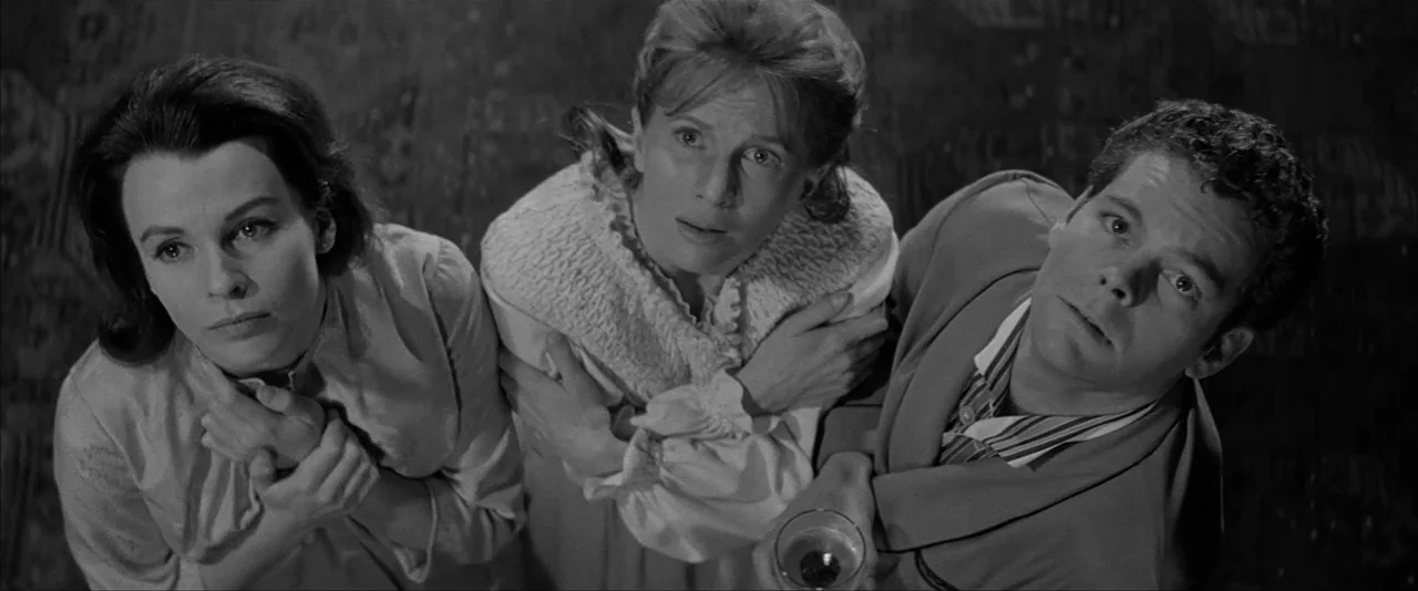 The Haunting (1963) movie still: medium — A dimly lit indoor space with textured walls and patterned flooring. Three people stand close together…