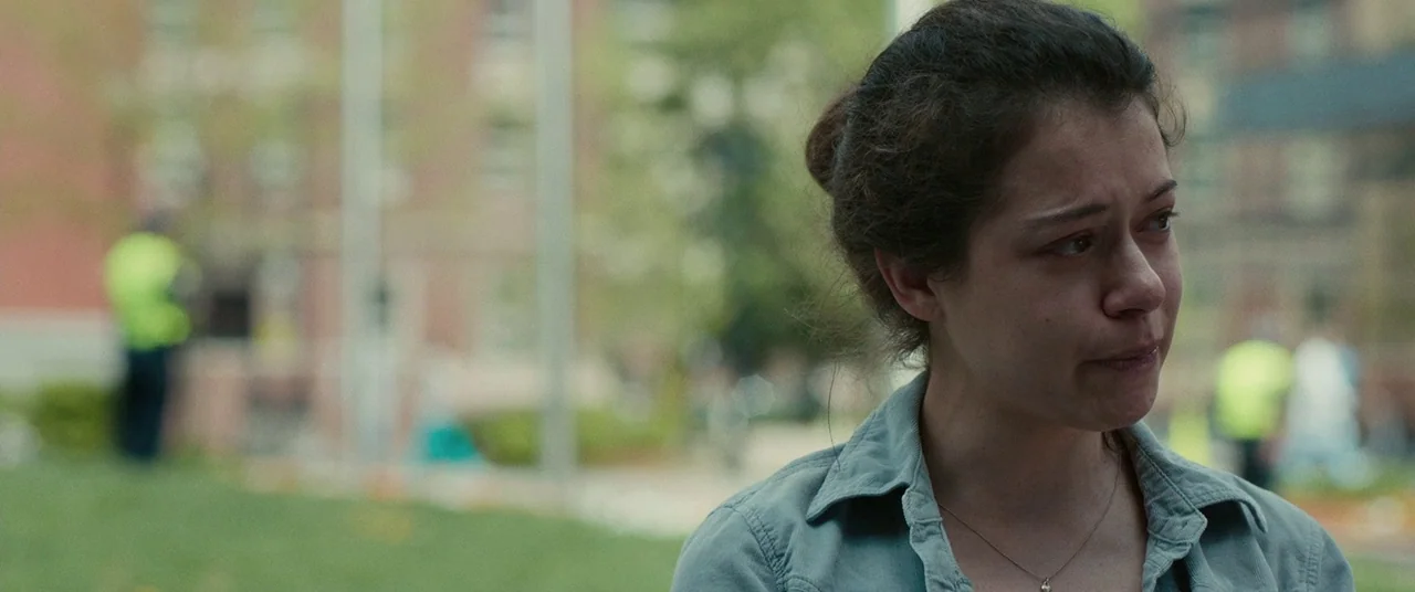 Stronger (2017) movie still: closeup — A blurred outdoor area with green lawns and brick buildings in the distance. A young woman with damp, red…