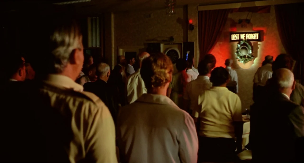 Wake In Fright (1971) movie still: medium — A dimly lit community hall features a wall with a red glowing memorial sign. A crowd of people stands in…