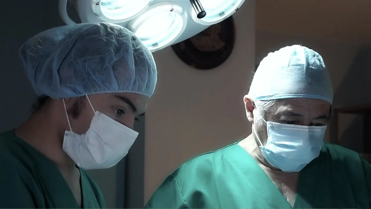 El Duende Sátiro (2016) movie still: medium — Two surgeons in scrubs stand closely together under bright overhead lighting. They focus downward…