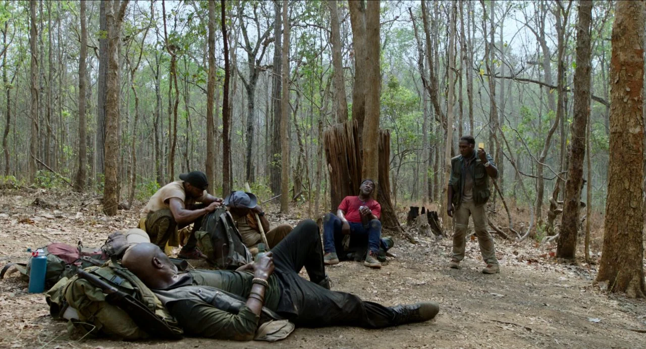 Da 5 Bloods (2020) movie still: wide — A group of men in tactical gear are resting within a sparse deciduous forest. Several backpacks and equipment…