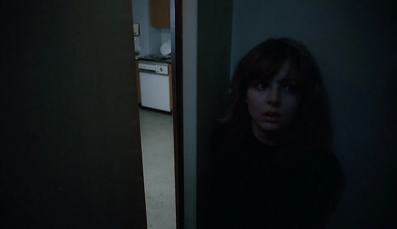 Angst (1983) movie still: medium — A dark apartment corridor with a narrow view into a brighter, lit domestic space. A young woman stands in the…