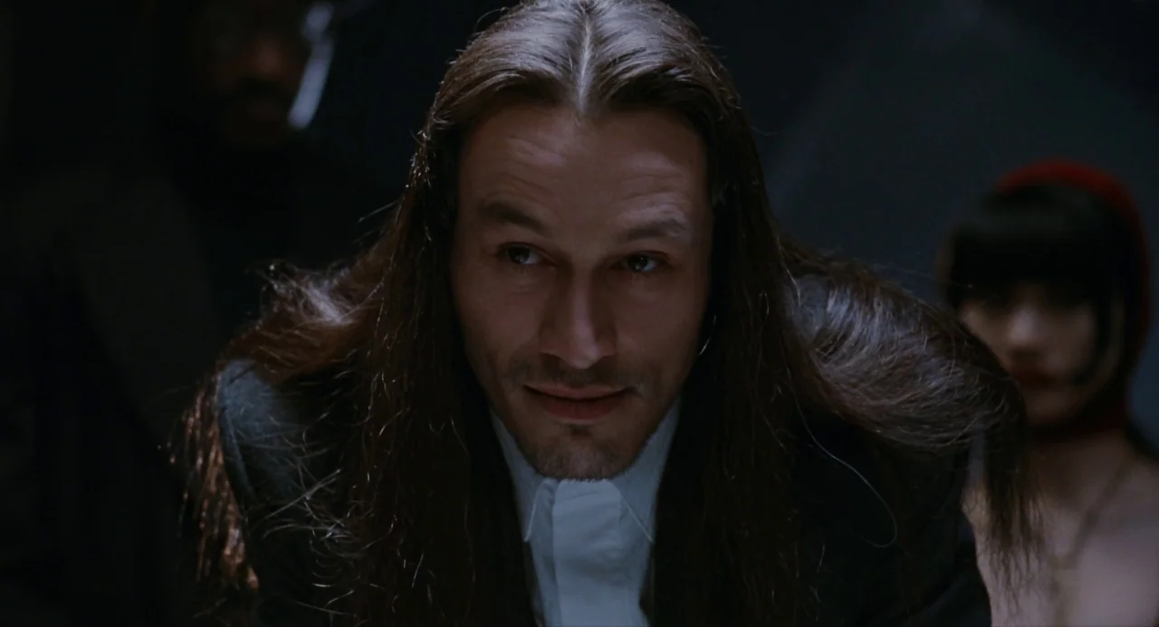 The Crow (1994) movie still: closeup — A dark, minimally lit space holds several people in the shadows. The central man with long hair stares…